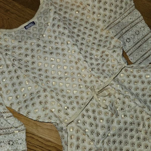 Beige klänning med paljetter från Amarillo - Säljer en superfin beige klänning köpt på Mall av India i New Delhi. Den har ett vackert paljettmönster som glittrar i ljuset och trekvartsärmar med spetsdetaljer. Klänningen har en knytning i midjan för en smickrande passform. Perfekt för festliga tillfällen eller en fin solig dag. 