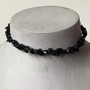 Snyggt svart choker-halsband med små, oregelbundna stenar som ger en cool och edgy look. Perfekt för att lyfta en outfit till fest eller vardag. Halsbandet har ett enkelt guldspänne som gör det lätt att ta på och av. Passar perfekt till både casual och mer uppklädda tillfällen.