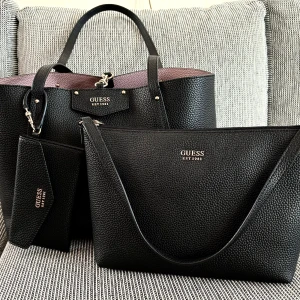 Guess bag -  Jättefin äkta Guess väska Brenton tote set modell med tillhörande mindre väska. Väskan gar att vända sa man kan välja om man vill ha den svarta eller lila sidan ut, sa egentligen blir det tre vaskor. Väska är endast använd två ganger. 