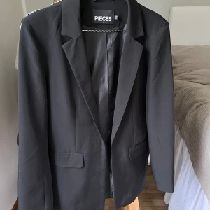 Svart kavaj från Pieces - Säljer en stilren svart kavaj från Pieces i storlek S. Perfekt för både vardag och fest! Kavajen har en klassisk design med krage och fickor, samt en slits baktill. Den är i nyskick skick och passar till många olika outfits✨