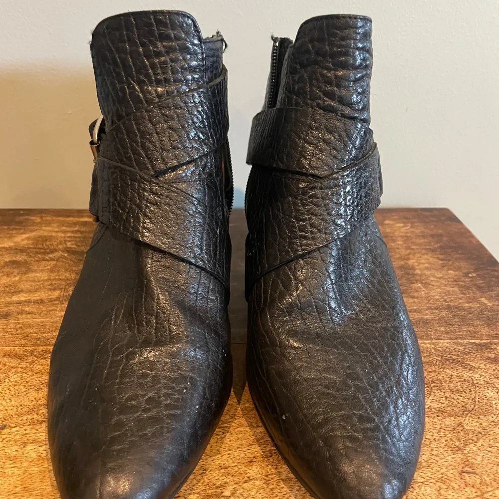 Snygga svarta boots i skinn med en cool spetsig tå och klack. De har en bred rem med ett stort metallspänne som ger en edgy look. Perfekta för höst/vår, och de passar både till vardags och fest. Skorna har en dragkedja på sidan. Fin ny sula.. Kengät.