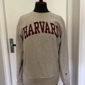 Beige Harvard sweatshirt från Champion - Säljer en vit sweatshirt från Champion med Harvard-tryck i vinrött på bröstet. Tröjan är långärmad och har en klassisk passform, perfekt för både chill och skola. Perfekt för hösten och vintern 
