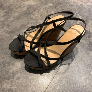 Svarta sandaler med kilklack - Snygga svarta sandaler med kilklack och remmar i mocka. Perfekta för sommarens alla fester! De har en bekväm passform och ger bra stöd med sina korsade remmar.