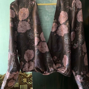 Blommig omlottopp i satin - Säljer en superfin blommig omlottopp i satin. Den har en djup v-ringning och långa ärmar. Perfekt för både fest och vardag! 🌸