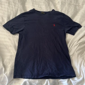 Mörkblå t-shirt från Polo Ralph Lauren - Säljer en snygg mörkblå t-shirt från Polo Ralph Lauren. Den är i storlek M och har en röd broderad logga på bröstet. T-shirten är i bomull och har korta ärmar. Perfekt för en avslappnad och stilren look!