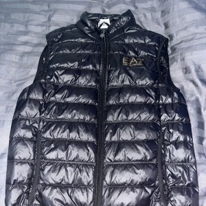 Svart dunväst från EA7 Emporio Armani - Säljer en snygg svart dunväst från EA7 Emporio Armani. Västen är quiltad och har en dragkedja framtill. Perfekt för höst och vinter, och den är i nyskick. Västen har en normal passform och är riktigt bekväm att ha på sig. Passar både till vardags och lite finare tillfällen. Storlek S.
