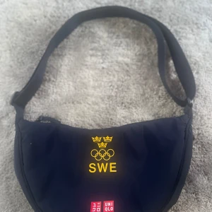 Blå axelväska från Uniqlo med SWE-tryck - Säljer en blå axelväska från sweden olympisk med ett gult SWE-tryck och olympiska ringar. Väskan har en justerbar axelrem och en dragkedja upptill. Perfekt för vardagsbruk eller som en sportig accessoar. OS VÄSKA