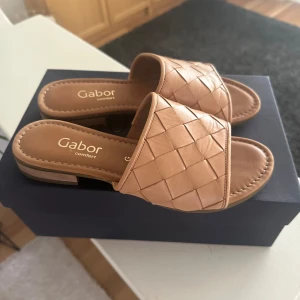 Beige sandaler från Gabor - Säljer ett par bekväma och stiliga sandaler från Gabor i beige färg. Sandalerna har en flätad design på ovandelen och är tillverkade i skinn. De har en låg klack och en mönstrad sula för bättre grepp. Perfekta för sommaren!
