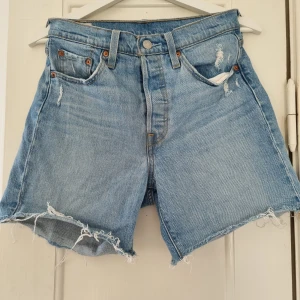 Blå jeansshorts från Levi's - Säljer ett par snygga blå jeansshorts från Levi's i modell 501. De är i nyskick och har en cool distressed look med slitna detaljer och fransiga kanter. Shortsen har en knappgylf och klassiska fem fickor. Perfekta för sommaren och kan stylas med allt från t-shirts till skjortor. Passa på att köpa dessa trendiga shorts! 💙 Omkrets midja: 68 cm, höft: 94 cm. 