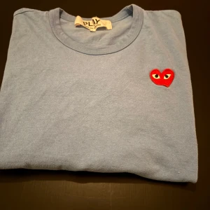 Blå t-shirt från Comme des Garçons - Snygg blå t-shirt från Comme des Garçons med det ikoniska röda hjärtat broderat på bröstet. Perfekt för en stilren och avslappnad look.