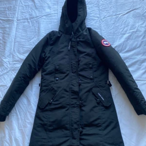 Svart parkajacka från Canada Goose - Säljer en svart parkajacka från Canada Goose med huva och flera praktiska fickor. Jackan har en stilren design med knappar och dragkedja framtill. Modell kensington parka men saknas päls är lite osäker av äkthet därav pris bra skick 