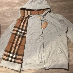 Säljer en stilren grå hoodie från Burberry med dragkedja och klassiskt rutigt mönster på insidan av huvan. Perfekt för en avslappnad look. Hoodien har ribbade muddar och justerbar dragsko i huvan.