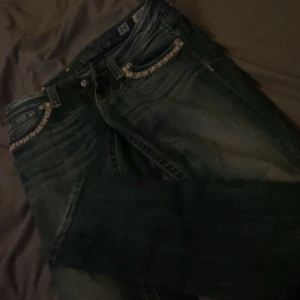 Jeans från Miss Me - Snygga jeans från Miss Me med dekorativa detaljer på fickorna och en mörk tvätt. Perfekta för dig som vill ha något unikt i garderoben. De har en klassisk femficksdesign och knappar med logotyp. Dom är skinny men ska sy om dom till bootcut