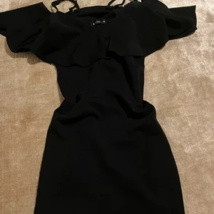 Svart klänning med volangdetalj - Elegant svart klänning med offshoulder-design och volangdetaljer. Perfekt för en stilren look.