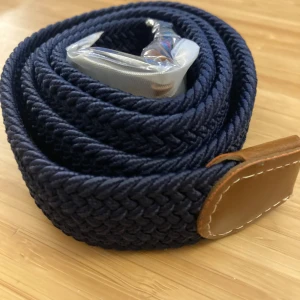 Mörkblått flätat bälte med bruna läderdetaljer (Grisch) - Snyggt mörkblått flätat bälte med stilrena bruna läderdetaljer och en silverfärgad spänne. Perfekt för att ge en klassisk touch till din outfit. 