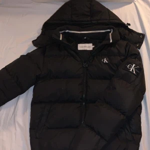 Svart dunjacka från Calvin Klein - Säljer en svart dunjacka från Calvin Klein med huva och dragkedja. Jackan har en broderad CK-logga på bröstet och är perfekt för kyliga dagar. Den är långärmad och har en stilren design.
