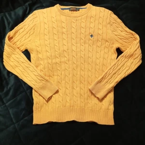 Orange Morris tröja - Säljer en snygg orange stickad tröja från Morris med kabelstickat mönster och en blå broderad detalj på bröstet. Tröjan har långa ärmar och ribbade muddar vid ärmslut och nederkant. Perfekt för en stilren look! Den är lite liten i storlek