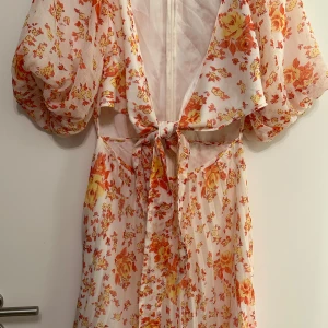Blommig klänning med puffärmar - Charmig blommig klänning i vitt med orange och gula blommor. Den har puffiga ärmar och en knytning i midjan. Perfekt för en somrig look! Klänningen är från ASOS design använd endast engång, st 38.
