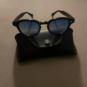 Moscot solglasögon - Snygga svarta solglasögon från Moscot med rund form och blåa linser. Perfekta för en stilren look. Kommer med ett matchande svart fodral. Kostar 4500kr nya, i nyskick men passar inte mitt ansikte. Storleken är 46mm vilket är average på denna modell då det finns 49mm som är large tex. 
