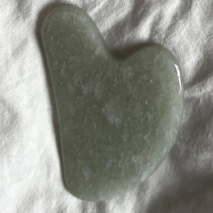 Grön gua sha-sten - Säljer en hjärtformad gua sha-sten i en vacker grön nyans. Perfekt för ansiktsmassage och hudvård. Stenen har en slät yta och är lätt att använda för att förbättra blodcirkulationen och minska svullnad.