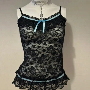 Svart spetslinne med blå detaljer - Elegant svart spetslinne med blommönster och ljusblå detaljer. Linnet har smala axelband och en söt rosett framtill. Perfekt för en stilfull look.