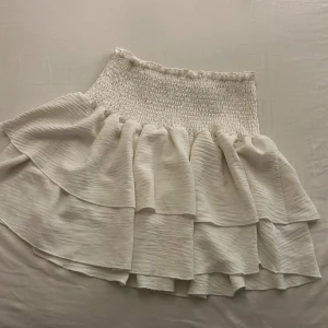 Vit volangkjol - Superfin vit kjol med volanger, perfekt för sommardagar! Den har en elastisk midja som gör den bekväm och lätt att bära. Kjolen är kort och har en luftig känsla, vilket gör den idealisk för både vardag och fest. Passar perfekt med en enkel topp eller blus.💕