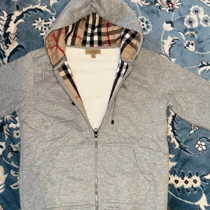Grå hoodie från Burberry - Säljer en snygg grå hoodie från Burberry i storlek L. Den har en klassisk dragkedja och en ikonisk rutig insida i huvan. Broderad logga på bröstet ger en exklusiv touch. Perfekt för både höst och vår! Tröjan är i nyskick och i storlek L men passar även M! Pris 999kr men går att diskutera! Möts upp i Stockholm eller fraktas
