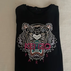Svart t-shirt med tigertryck från Kenzo - Säljer en svart t-shirt från Kenzo med ett coolt tigertryck i vitt och rosa på framsidan. Perfekt för att ge din outfit en edgy touch. T-shirten är i bra skick och passar perfekt till både vardag och fest. 🐯✨