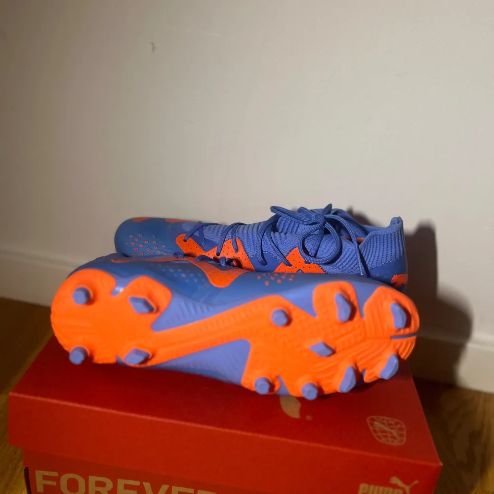 Säljer ett par snygga fotbollsskor från Puma i blått och orange. Skorna har en cool design med snörning och är perfekta för fotbollsträning eller match. De är i nytt skick och kommer i originalkartong. Perfekt för dig som vill sticka ut på planen! ⚽️. Skor.