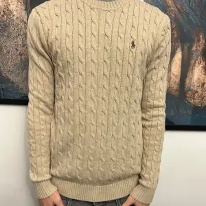 Säljer en snygg beige stickad tröja från Ralph Lauren. Den har ett klassiskt kabelstickat mönster och en liten broderad logga på bröstet. Perfekt för höst och vinter, med långa ärmar och ribbade muddar. Passar både till vardags och lite finare tillfällen.