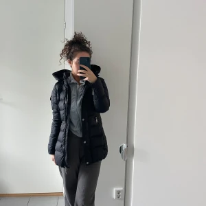 Svart vinterjacka från Rockandblue - Snygg svart  vinterjacka från Rockandblue med huva och dragkedja. Jackan har en figursydd passform och detaljer som en liten ficka med dragkedja på ärmen. Perfekt för kyligare dagar. Jackan har använts ett väldigt fåtal gånger - så mycket bra skick.