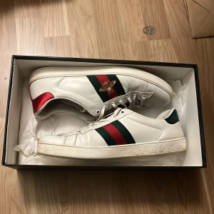 Vita sneakers från Gucci - Säljer ett par vita Gucci sneakers storlek 46  med gröna och röda ränder samt en broderad insekt på sidan. Skorna har en röd och en grön hälflik med Gucci-logga. Perfekta för en stilren look.