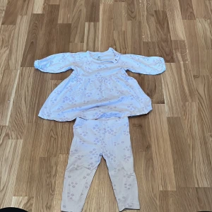 Vit och lila blommig klänning med leggings från ICA - Söt vit klänning med lila blommönster och matchande leggings från ICA. Tillverkad i ekologisk bomull, perfekt för små barn. Klänningen har långa ärmar och en bekväm passform.