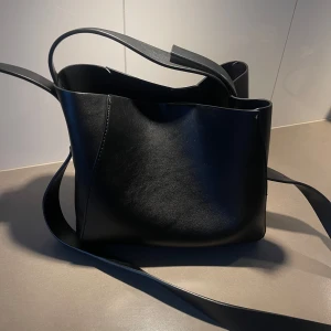 Svart axelväska i skinn - Snygg och stilren svart axelväska i skinn med en praktisk dragkedja inuti. Perfekt för att bära dina dagliga nödvändigheter med stil. Väskan har en enkel design med en bred axelrem för bekväm användning.