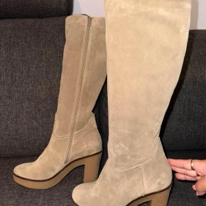 Beige mockastövlar med klack - Snygga beige stövlar i mocka med hög klack och dragkedja på sidan. Perfekta för höst och vinter, dessa stövlar ger en elegant touch till vilken outfit som helst. De är i mycket bra skick och redo att användas för både vardag och fest.