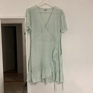 Ljusgrön blommig omlottklänning - Charmig ljusgrön omlottklänning med blommigt mönster och volangdetaljer. Klänningen har korta ärmar och knytband i midjan för en smickrande passform. Perfekt för somriga dagar!