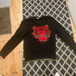 Svart tröja från Kenzo med tigerbroderi - Säljer en svart tröja från Kenzo i storlek XL. Den har ett coolt tigerbroderi i rött och guld på framsidan. Perfekt för höst och vinter med sin långärmade design. Tröjan är i mycket bra skick och redo för nya äventyr! 🐯✨ pris kan diskuteras