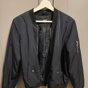 Mörkblå bomberjacka från ONLY - Snygg mörkblå bomberjacka från ONLY med guldig dragkedja och ribbade muddar. Jackan har en klassisk bomberstil med fickor fram och en ärmficka med dragkedja. 