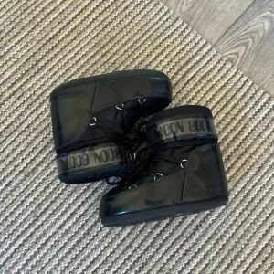 Svarta Moon Boots - Säljer ett par svarta Moon Boots med hög glans och snörning. Perfekta för vintern med sin unika design och varma passform. De har en tydlig logotyp runt skaftet och är idealiska för kalla dagar. Modellen är icon low och är inköpta för 2200kr på zalando, säljer eftersom de aldrig används