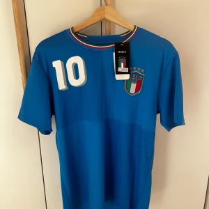 Blå fotbollströja från Italien - Snygg blå fotbollströja från Italien med nummer 10 och R. Baggio på ryggen. Tröjan har korta ärmar och ett stilrent märke på bröstet. Perfekt för fotbollsfans!