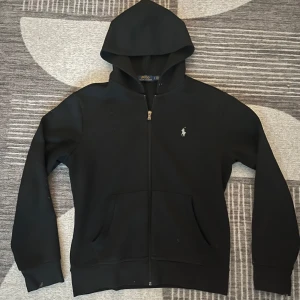 Svart hoodie från Polo Ralph Lauren - Säljer en klassisk svart hoodie från Polo Ralph Lauren med dragkedja och broderad logga på bröstet. Perfekt för en stilren och bekväm look. 🖤