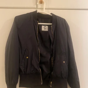 Svart bomberjacka från Cubus - Säljer en snygg svart bomberjacka från märket C i storlek 164. Jackan har en dragkedja framtill och två fickor med knappar. Perfekt för höst och vår, med ribbade muddar vid ärmslut och nederkant. Passar både till vardags och fest!
