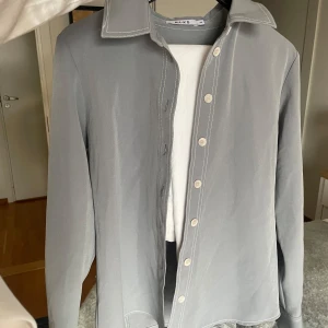 Grå skjorta från NA-KD - Säljer en oanvänd stilren oversized grå skjorta från NA-KD i storlek S. Skjortan är tillverkad av 95% polyester och 5% elastan, vilket ger den en skön passform. Den har långa ärmar och knappar framtill. 