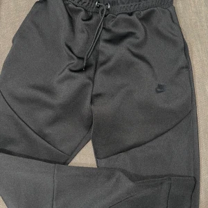 svarta Nike Tech byxor från Nike - säljer ett par svarta Nike Tech byxor från Nike i mycket bra skick. de har en elastisk midja med snörning och praktiska dragkedjor vid fickorna. de är perfekta för både träning och chill dag. (OBS!! ganska små i strl, passar XS/S)