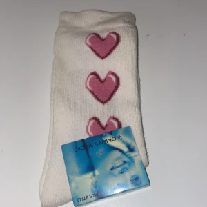 Vita strumpor med rosa hjärtan - Säljer ett par vita strumpor med tre söta rosa hjärtan på framsidan. De är i nytt skick och perfekta för att ge en gullig touch till din outfit. Strumporna är mjuka och bekväma, idealiska för vardagsbruk.