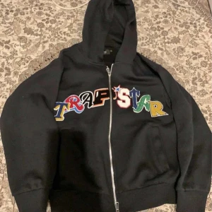 Svart hoodie med färgglatt tryck från Trapstar - Säljer en svart hoodie från Trapstar med ett coolt, färgglatt tryck över bröstet. Den har en dragkedja framtill och en skön huva. Perfekt för en avslappnad stil eller streetwear-look. Passar både höst och vår!