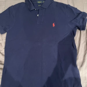 Mörkblå pikétröja från Polo - Säljer en snygg mörkblå pikétröja från Polo i storlek M. Tröjan har en klassisk krage med två knappar och det ikoniska röda Polo-logotypen broderad på bröstet. Perfekt för både vardag och fest! 💙