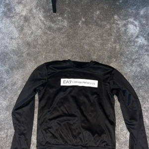 Svart långärmad tröja från EA7 Emporio Armani - Säljer en stilren svart långärmad tröja från EA7 Emporio Armani. Tröjan har en vit logga på bröstet och en mindre logga på baksidan vid nacken. Perfekt för en casual look eller att klä upp med accessoarer. Passar både till vardags och fest!
