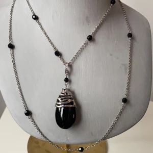 Silverfärgat halsband med svart hänge - Snyggt silverfärgat halsband med ett stort svart hänge. Kedjan är dekorerad med små svarta pärlor och har en justerbar längd. Hänget har en unik design med en silverfärgad infattning som ger en elegant touch. Perfekt för både vardag och fest!
