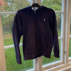 Mörkblå hoodie från Ralph Lauren - Säljer en mörkblå hoodie från Ralph Lauren i storlek S. Den har en dragkedja framtill och den klassiska loggan broderad på bröstet. Perfekt för höst och vår! Observera att det finns några små fläckar på framsidan som lätt går bort med lite behandling. Passar både till vardags och chill.